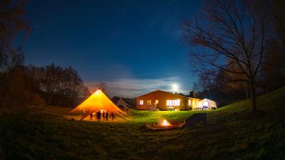 Oplyst hytte, bål og telt i skov med mørkeblå nattehimmel